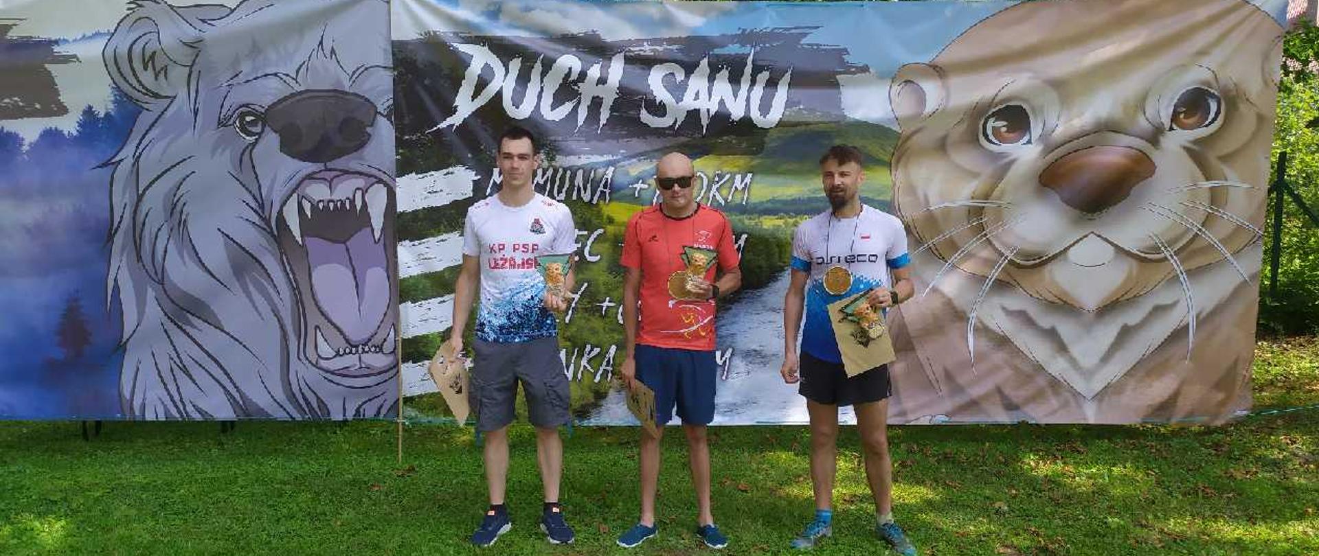 Na zdjęciu widzimy zwycięzców biegu z pucharami, w tym strażak z naszej komendy, który zajął 2 miejsce. W tle widzimy baner z logiem biegu.