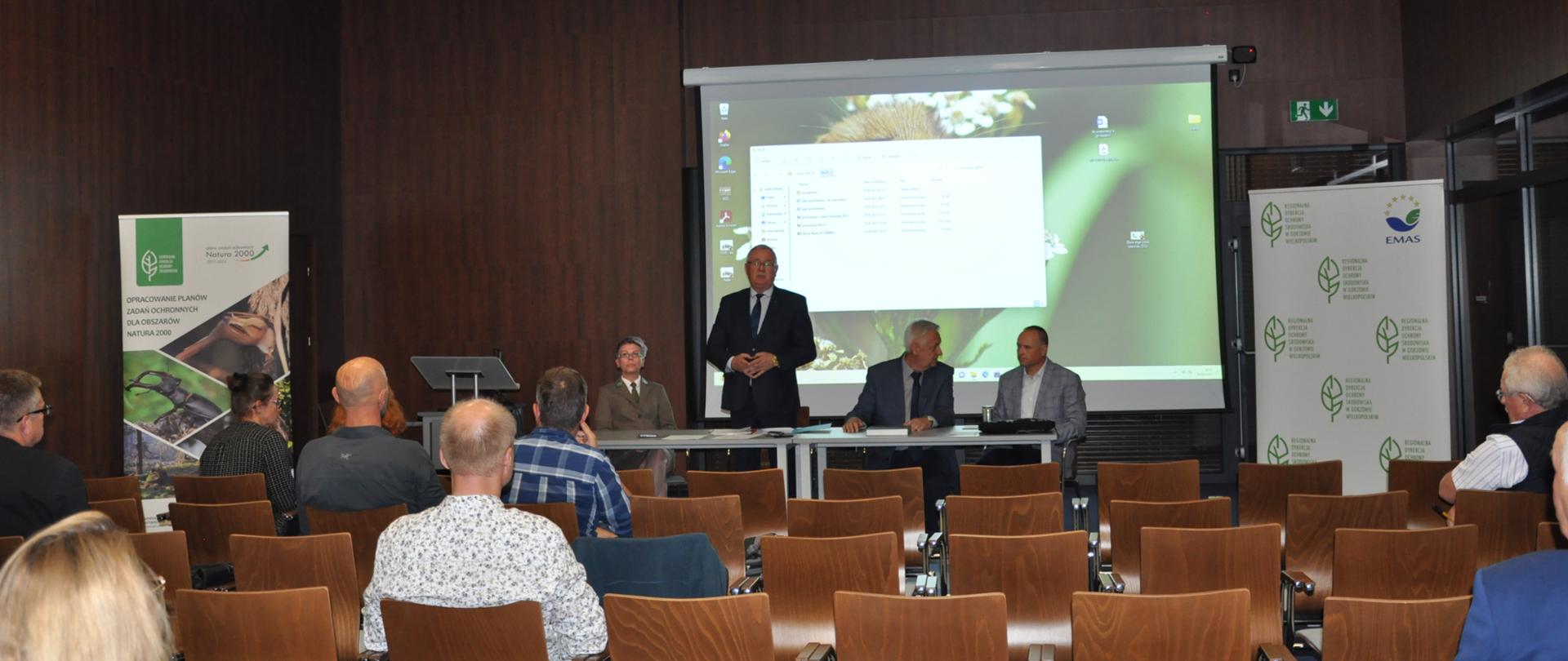 Na drugim planie za stołem siedzą cztery osoby. Przed nimi na krzesłach w dużej sali siedzi kilka osób.