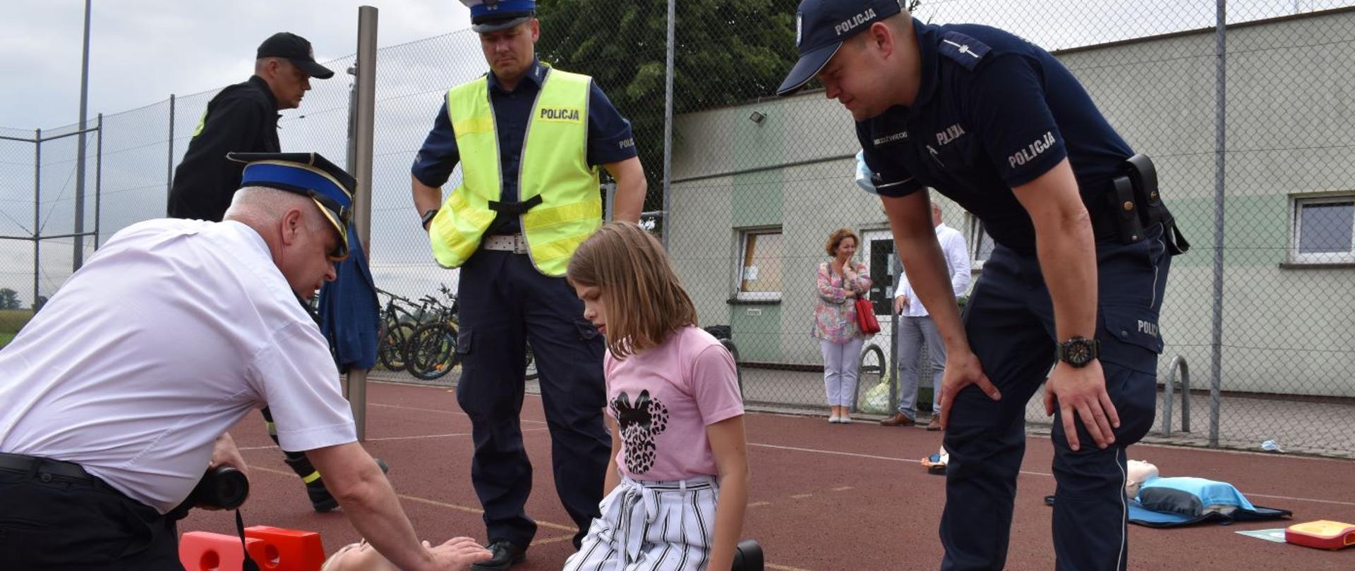 Boisko Zespołu Szkolno- Przedszkolnego w Zielonej Wsi. Na noszach - desce leży uczennica szkoły. Obok niej z jednej strony klęczy inna dziewczynka, z drugiej zaś - strażak, który prawą dłonią uciska klatkę piersiową leżącej. W ten sposób instruuje w jaki sposób prawidłowo prowadzić resuscytację krążeniowo-oddechową. Przyglądają się temu dwaj policjanci. 