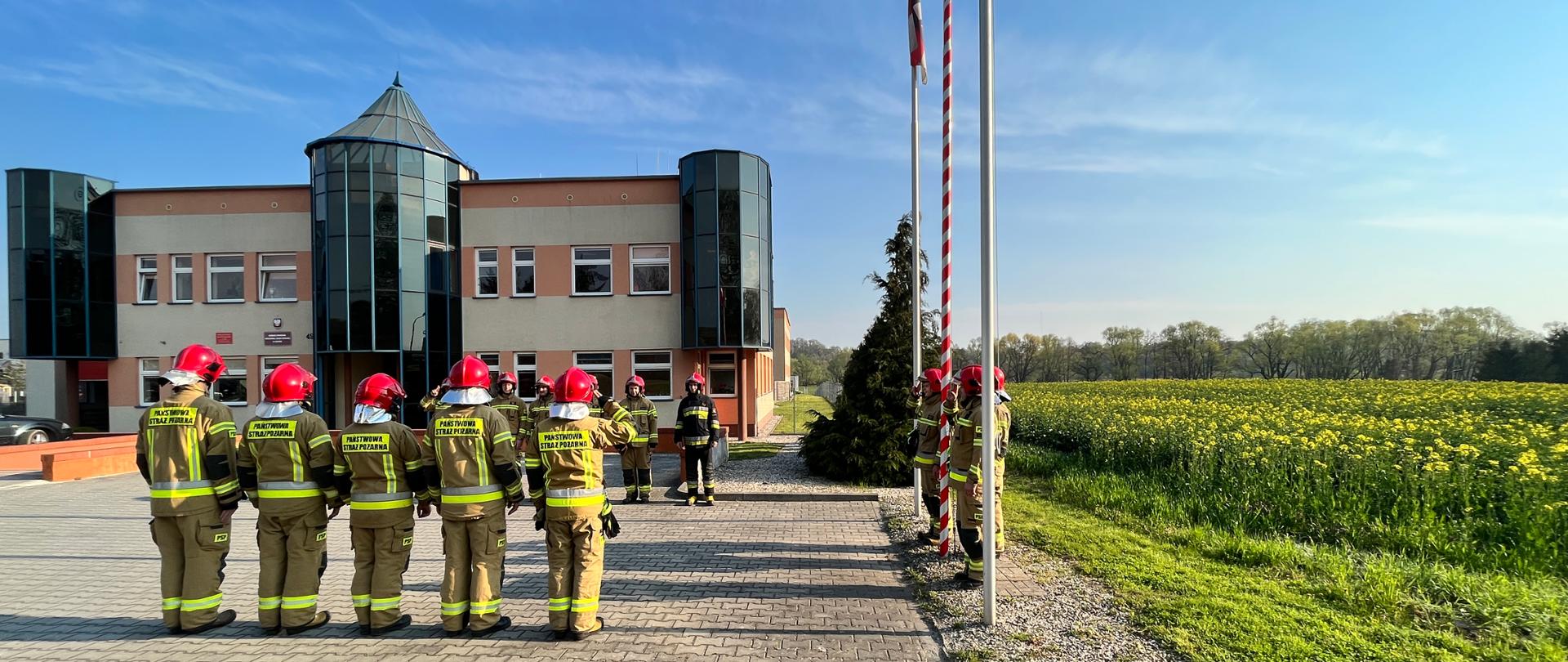 umundurowani strażacy podczas wciągania flagi państwowej na maszt, stojący na placu przed budynkiem komendy