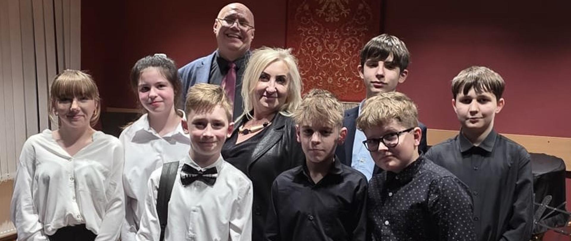 Pamiątkowe zdjęcie grupy młodych muzyków i dwojga opiekunów stojących na scenie po zakończonym konkursie. Uczniowie, ubrani w stroje galowe, trzymają kolorowe torby z upominkami, a w tle widać godło Polski na czerwonej ścianie.