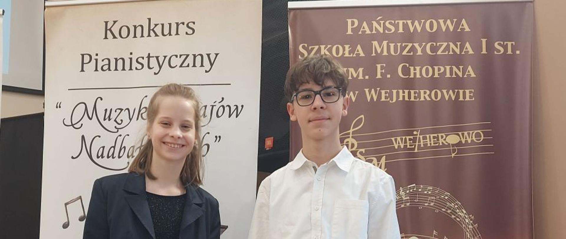 Zdjęcie kolorowe. Dwoje nastolatków stojących obok siebie między dwoma wysokimi, pionowymi banerami reklamowymi. Dziewczyna po lewej stronie ubrana w czarny, błyszczący strój i ciemną marynarkę, uśmiecha się. Chłopak po prawej stronie w białej koszuli z długim rękawem, granatowych jeansach i okularach na nosie, stoi prosto. Banery z napisami "Konkurs Pianistyczny 'Muzyka Krajów Nadbałtyckich'" i "PAŃSTWOWA SZKOŁA MUZYCZNA I ST. IM. F. CHOPINA W WEJHEROWIE" stanowią tło.