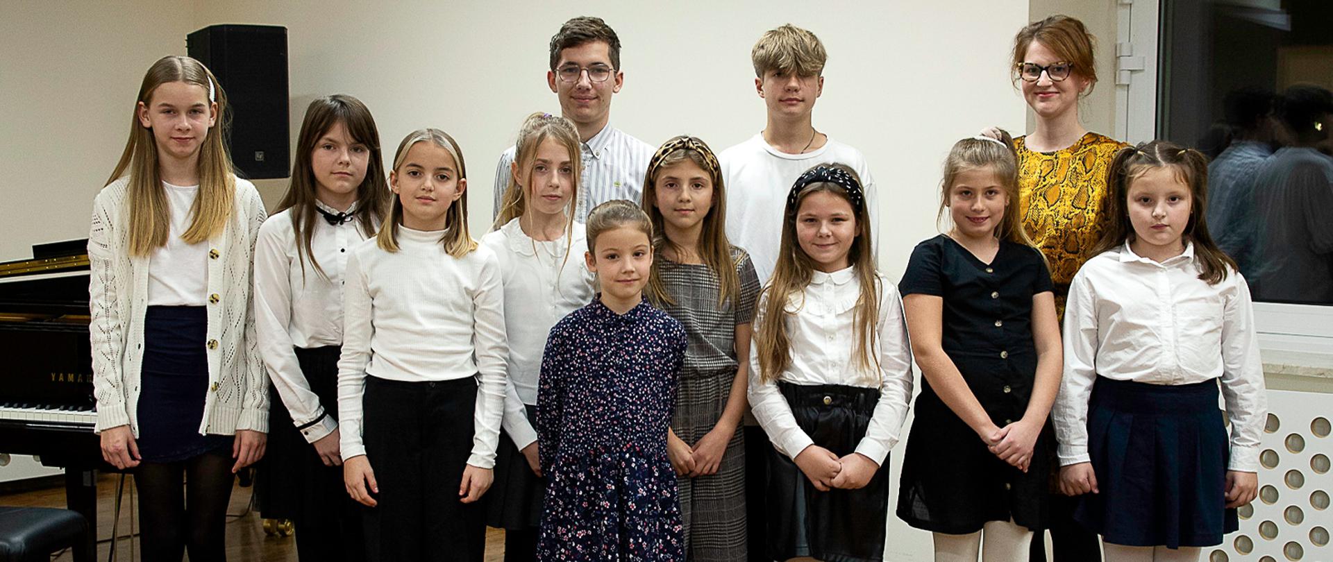Na zdjęciu widać grupę stojących osób, z przodu stoi 9 dziewczynek, za nimi dwóch chłopców i nauczycielka, wszyscy patrzą w kierunku robiącego zdjęcie. Z nimi po lewej stronie stoi widoczny częściowo fortepian, dalej jasna ściana z oknem.