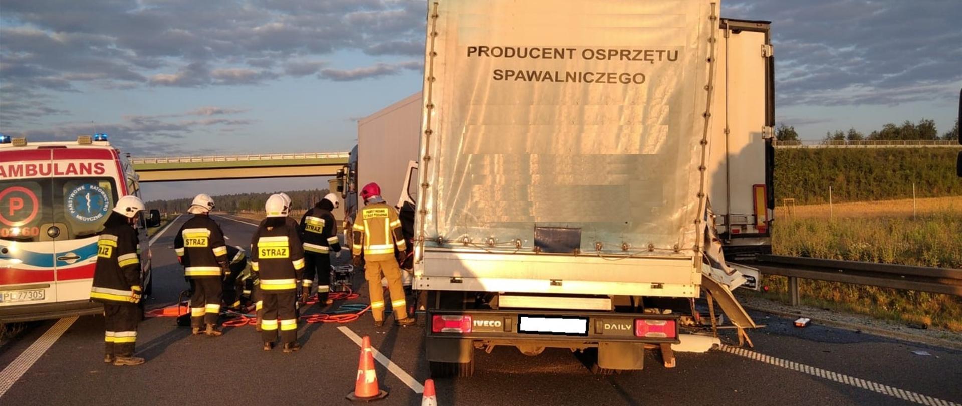 Widoczne dwa pojazdy ciężarowe biorące udział w wypadku oraz karetka pogotowia ratunkowego. Strażacy wykorzystując sprzęt hydrauliczny wykonują dostęp do kabiny pojazdu.