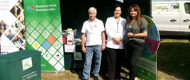 Mężczyzna i dwie kobiety pozują, stojąc na trawie na tle zielonego namiotu KRUS z napisem PRACUJ BEZPIECZNIE. Za nimi stolik z ulotkami i nagrodami konkursowymi.