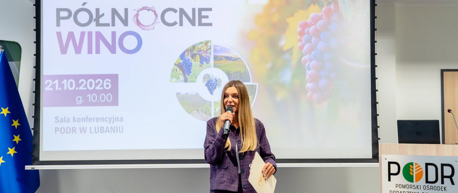 Kobieta w fioletowym garniturze przemawia do mikrofonu na tle ekranu z napisem: Północne Wino". 