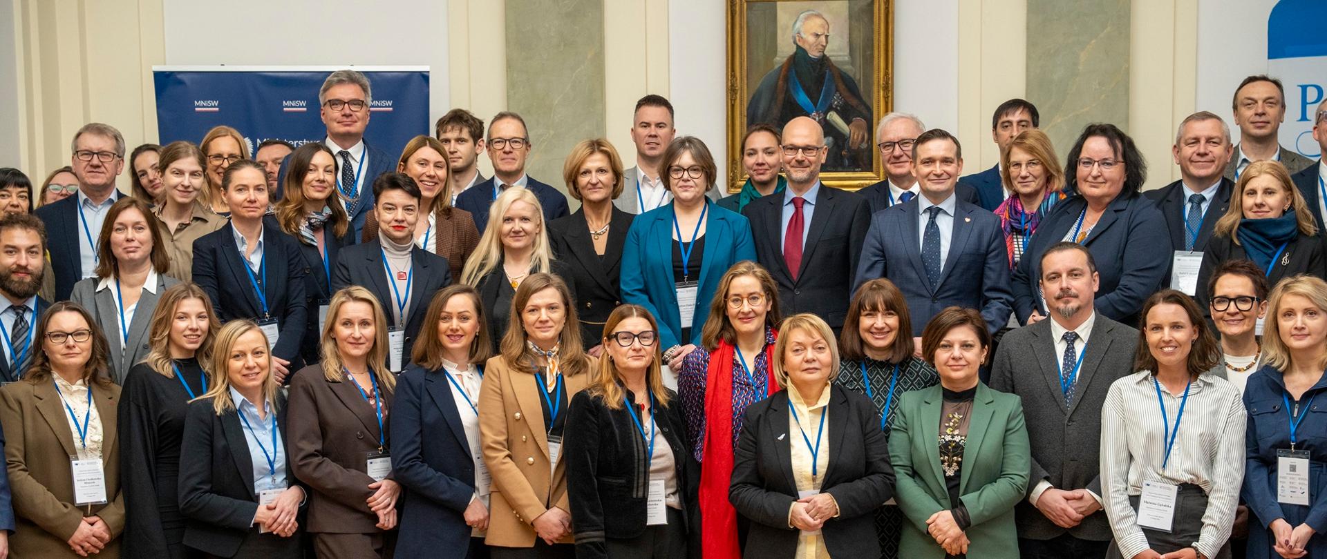 Zdjęcie zbiorowe, pod ścianą na której wisi portret stoi grupa kilkudziesięciu osób, pośrodku grupy wiceminister Zioło-Pużuk.