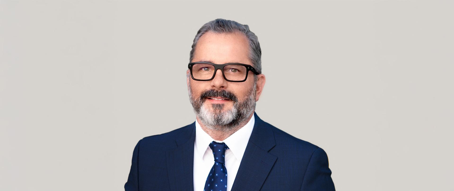 Mężczyzna z brodą w okularach w ciemnogranatowej marynarce, białej koszuli z ciemnoniebieskim krawatem lekko uśmiecha się do zdjęcia.