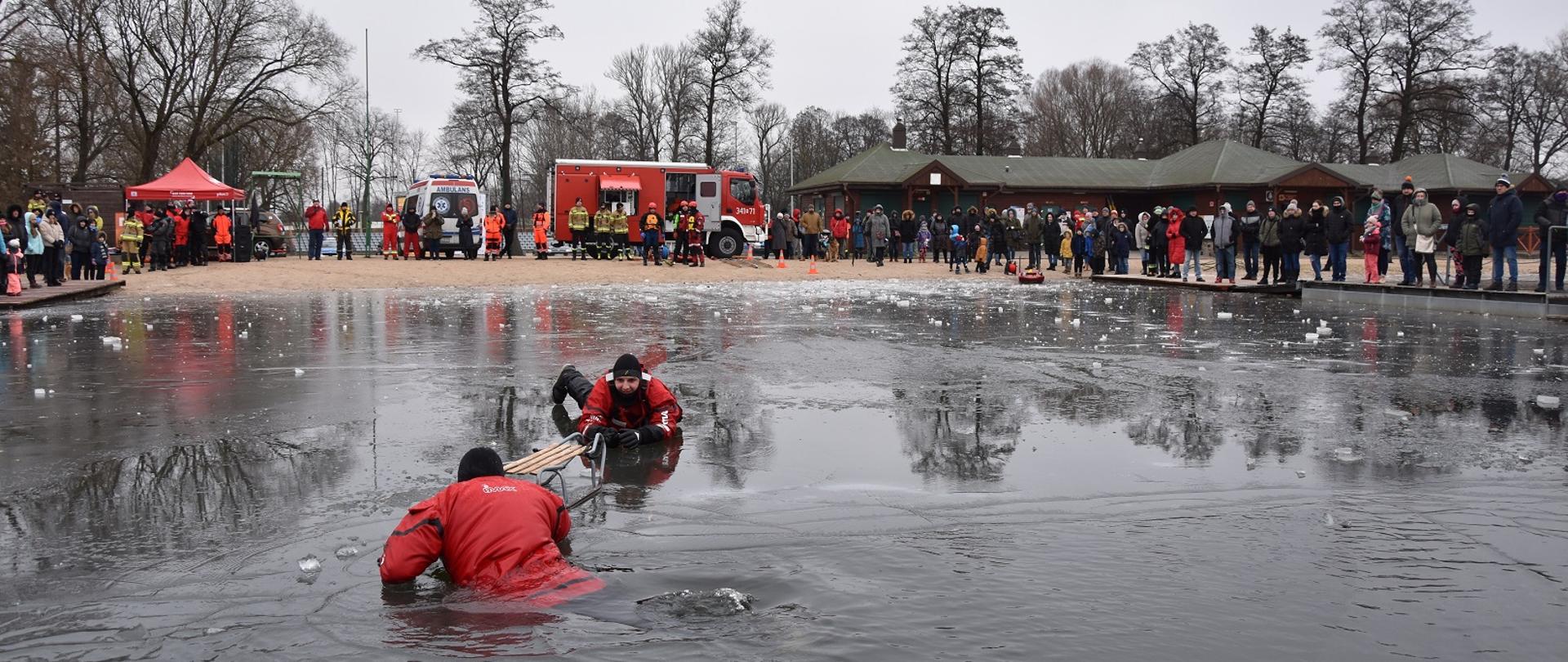Zdjęcie obrazuje osobę w wodzie a druga będąc na lodzie ją ratuje podając sanki w tle ludzie i samochody 