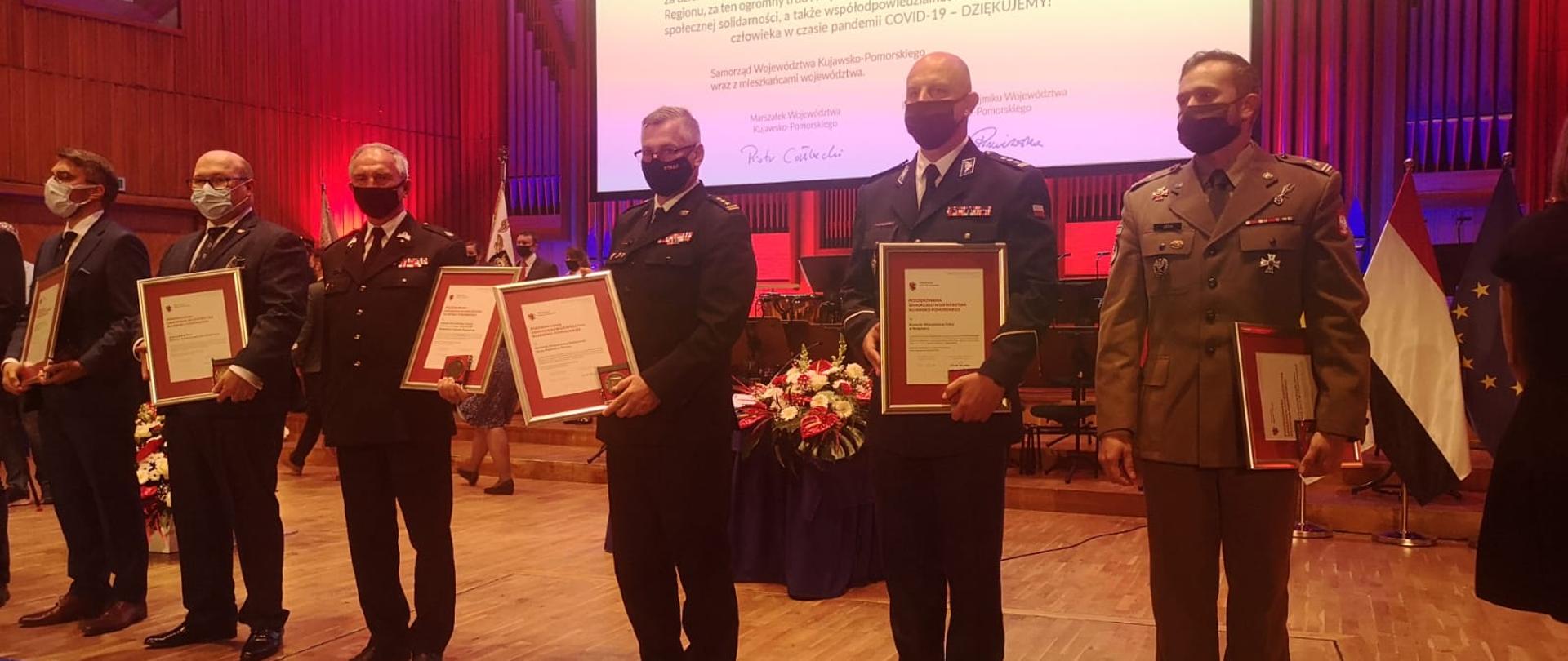 Na scenie Filharmonii Pomorskiej w Bydgoszczy stoją osoby, które odebrały tego dnia podziękowania za współdziałanie w pandemii. Wśród nich jest komendant wojewódzki PSP. Każda z osób, a widocznych jest 6, stoją w szeregu. Każdy z nich trzyma w ręku oprawiony dokument podziękowania oraz medal okolicznościowy - grawerton. W tle, na ścianie wyświetlony tekst podziękowań, widać także kwiaty, jako dekorację sceny.