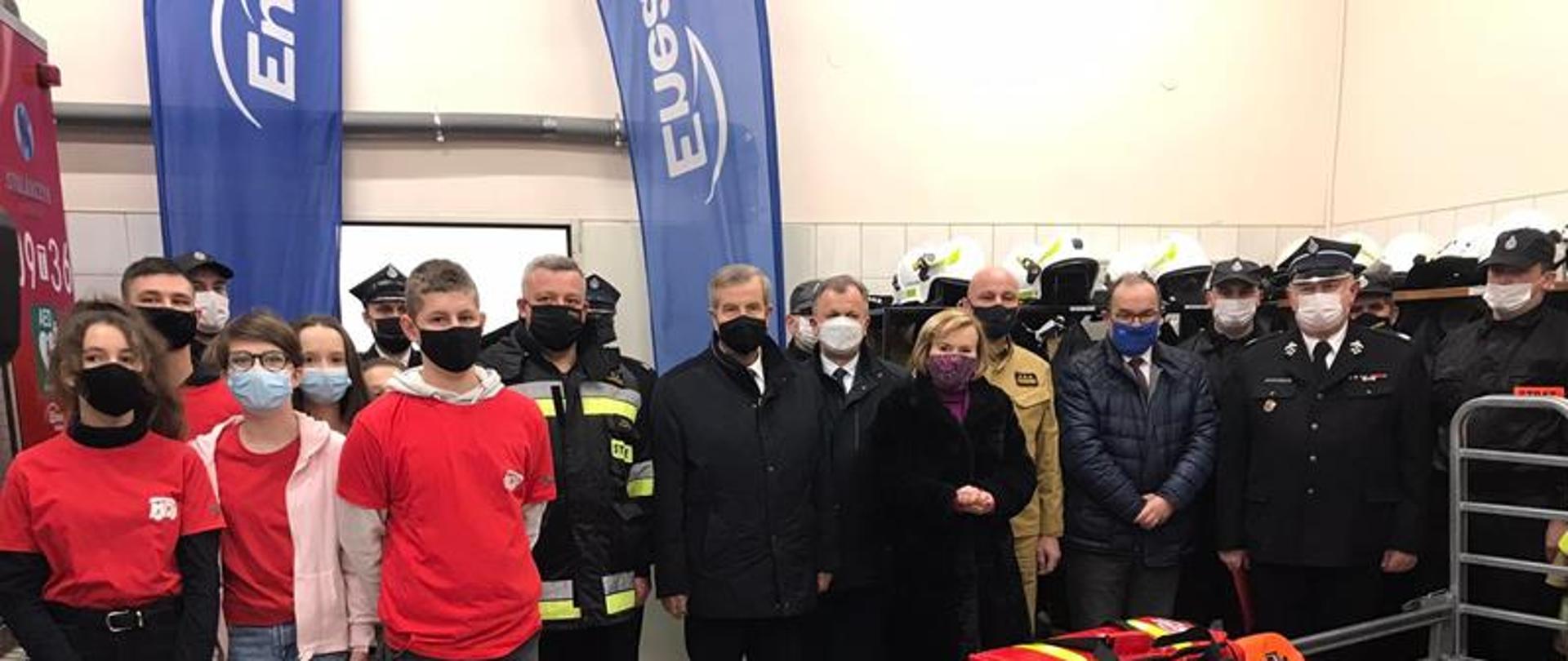 Funkcjonariusze, druhowie z OSP Połaniec oraz zaproszeni goście stoją obok nowego przekazanego sprzętu dla jednostki OSP Połaniec. Wszyscy maja założone maseczki. 