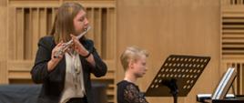 Przed pulpitem, na którym leżą nuty stoi młoda dziewczyna. Gra na flecie. Przy fortepianie akompaniuje jej kobieta.