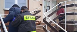 Strażak ochotniczej straży pożarnej pomaga starszej osobie wejść po schodach do przychodni, gdzie szczepi się osoby przeciwko COVID-19. W tle widać starszą panią, która czeka na mężczyzn.