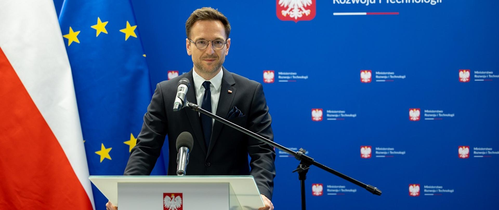 Minister rozwoju i technologii Waldemar Buda na konferencji prasowej, minister stoi za mównicą z mikrofonem, za jego placami flagi Polski i UE oraz niebieski baner z logo MRiT.