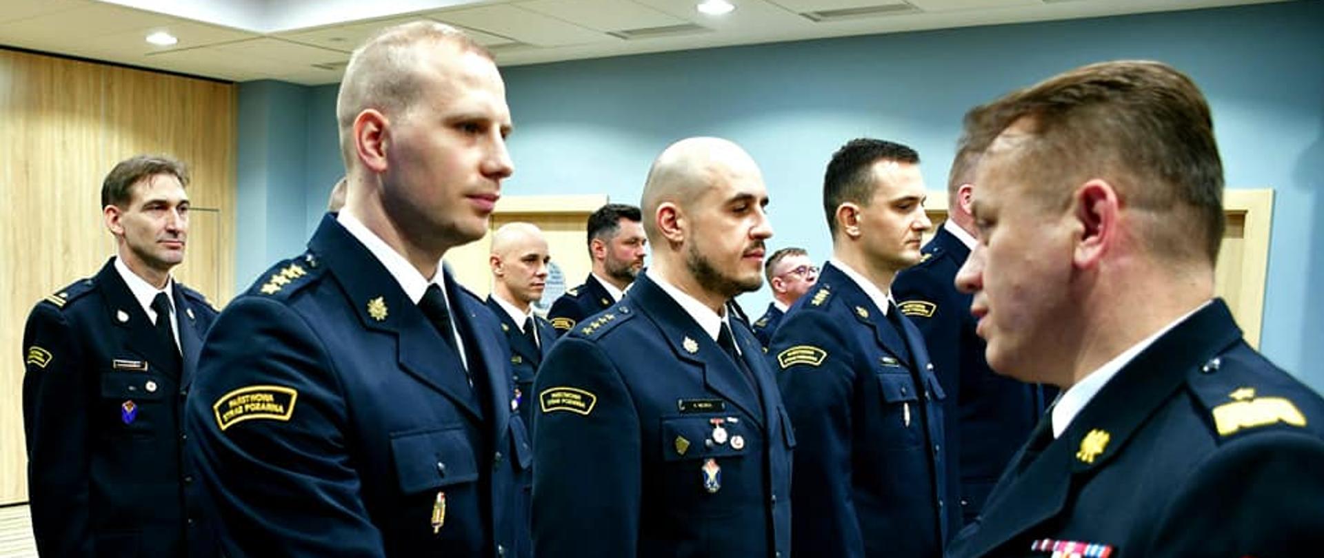 Mazowiecki Komendant Wojewódzki wręcza teczkę oficerowi Straży Pożarnej stojącym w jednym z dwóch szeregów.