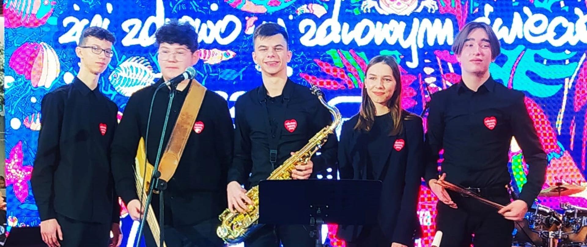 zdjęcie przedstawia członków zespołu kameralnego w składzie pianino, gitara basowa, saksofon, wokal, perkusja, w tle widać barwną grafikę 31. finał WOŚP