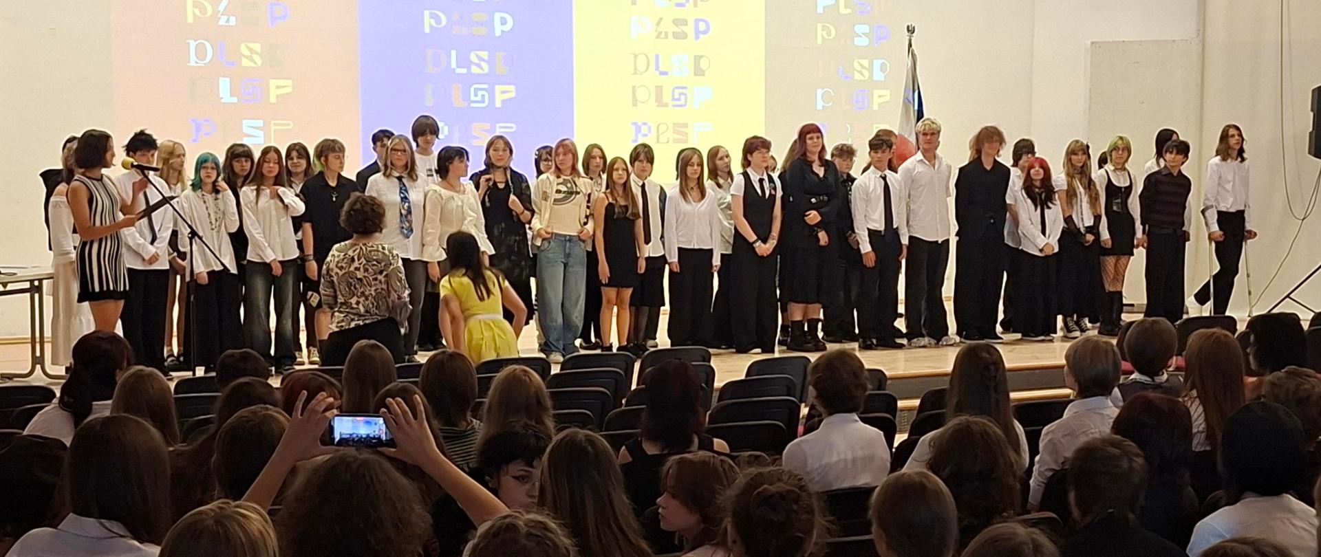 Uczniowie stoją na scenie szkolnej auli podczas uroczystości rozpoczęcia roku szkolnego. Przed nimi znajduje się publiczność, a w tle widoczna jest kolorowa projekcja graficzna oraz szkolny sztandar. Na scenie trwa prezentacja grupy uczniów.
