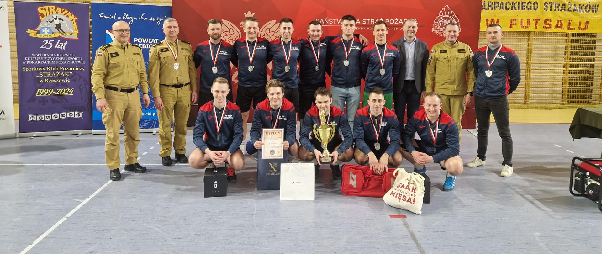 Reprezentacja KP PSP Ropczyce po dekoracji strazacy w dresach na tle baneru PSP