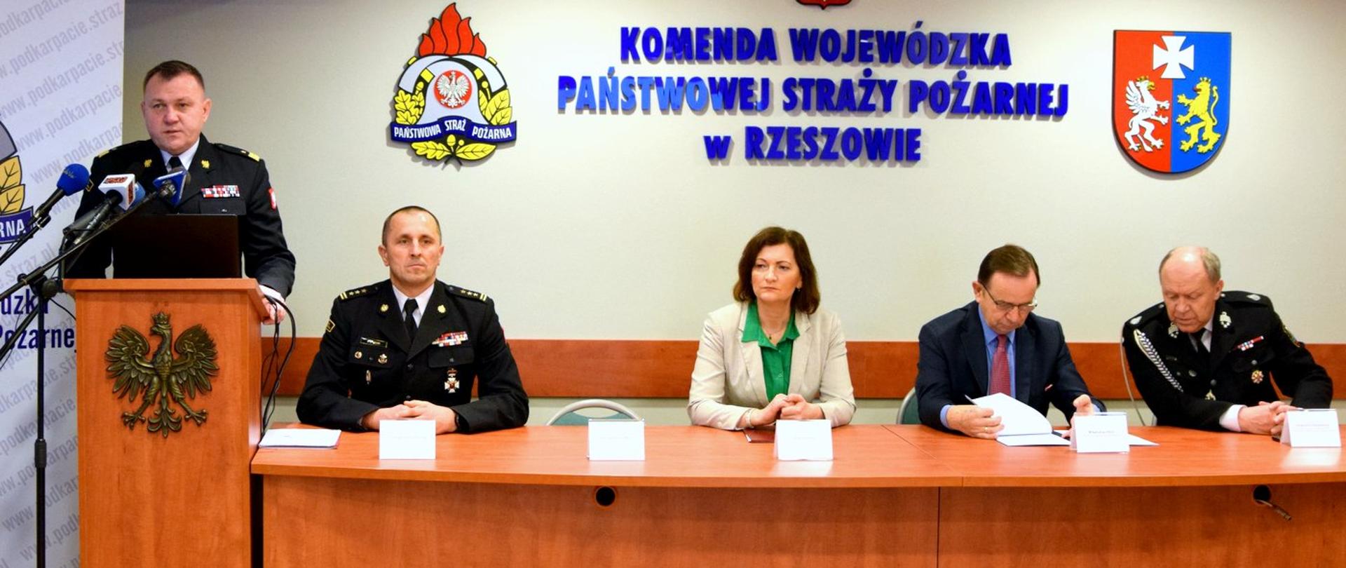 Zdjęcie zrobione w pomieszczeniu, podczas konferencji prasowej. Przy mównicy stoi oficer Państwowej Straży Pożarnej w stopniu generała. Przy stole konferencyjnym siedzą 4 osoby, wśród nich oficer Państwowej Straży Pożarnej w stopniu starszego brygadiera. Na ścianie, za uczestnikami konferencji, znajduje się napis Komenda Wojewódzka Państwowej Straży Pożarnej w Rzeszowie oraz logotypy Państwowej Straży Pożarnej i województwa podkarpackiego.