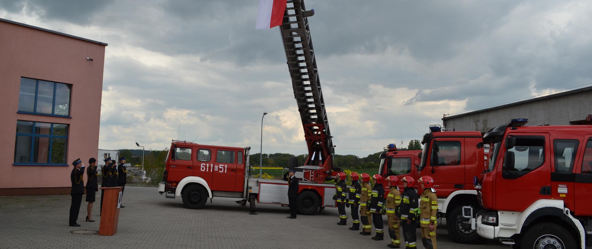 cztery samochody strażackie, przed nimi szereg strażaków salutujących do wciągniętej flagi państwowej