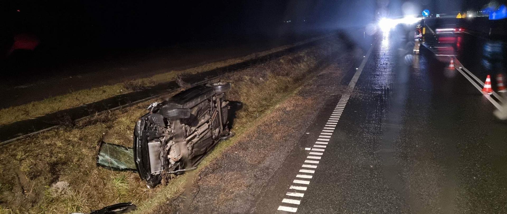 Samochód osobowy leży na prawym boku w przydrożnym rowie. 