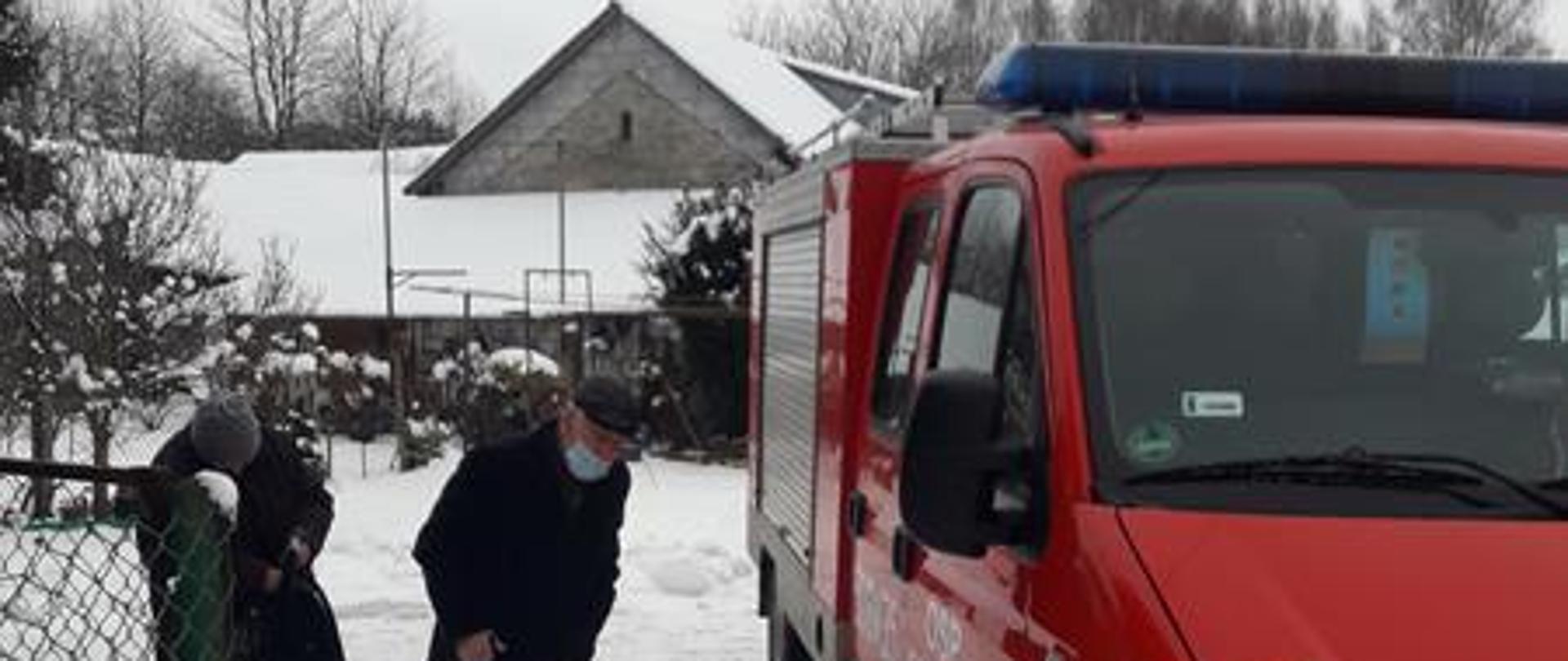 Widać dwie starsze osoby które przygotowują się żeby wsiąść do samochodu pożarniczego.