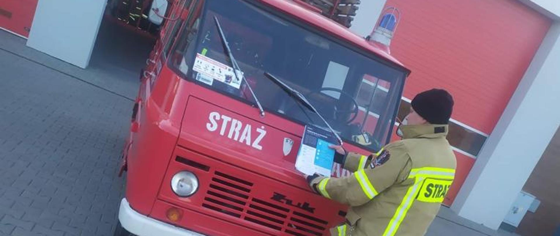 Na zdjęciu stoi strażak przy samochodzie strażackim. Jest obrócony tyłem a w ręcach trzyma ulotkę dotyczącą szczepień przeciwko COVID 19
