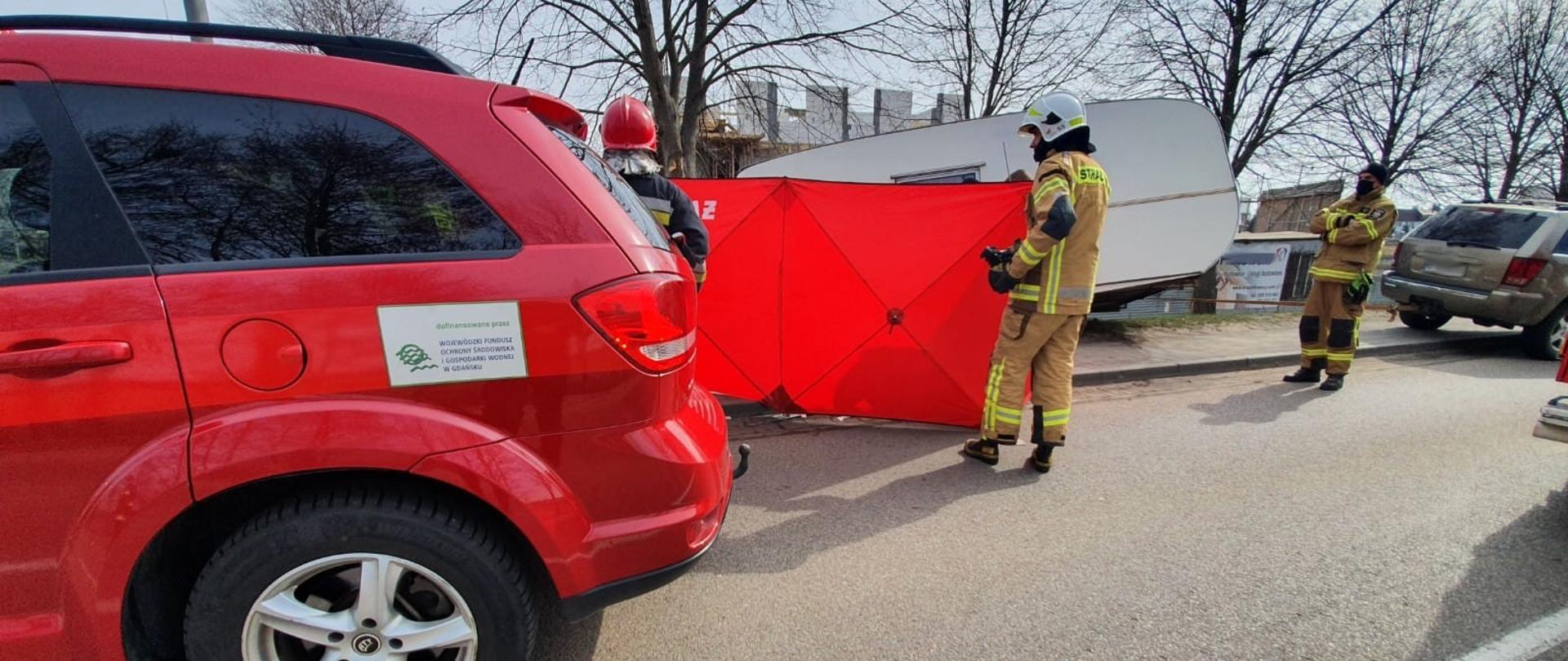 Na pierwszym planie, tył samochodu strażackiego, w tle rozwinięty parawan ochronny, w oddali odczepiona przyczepa kempingowa