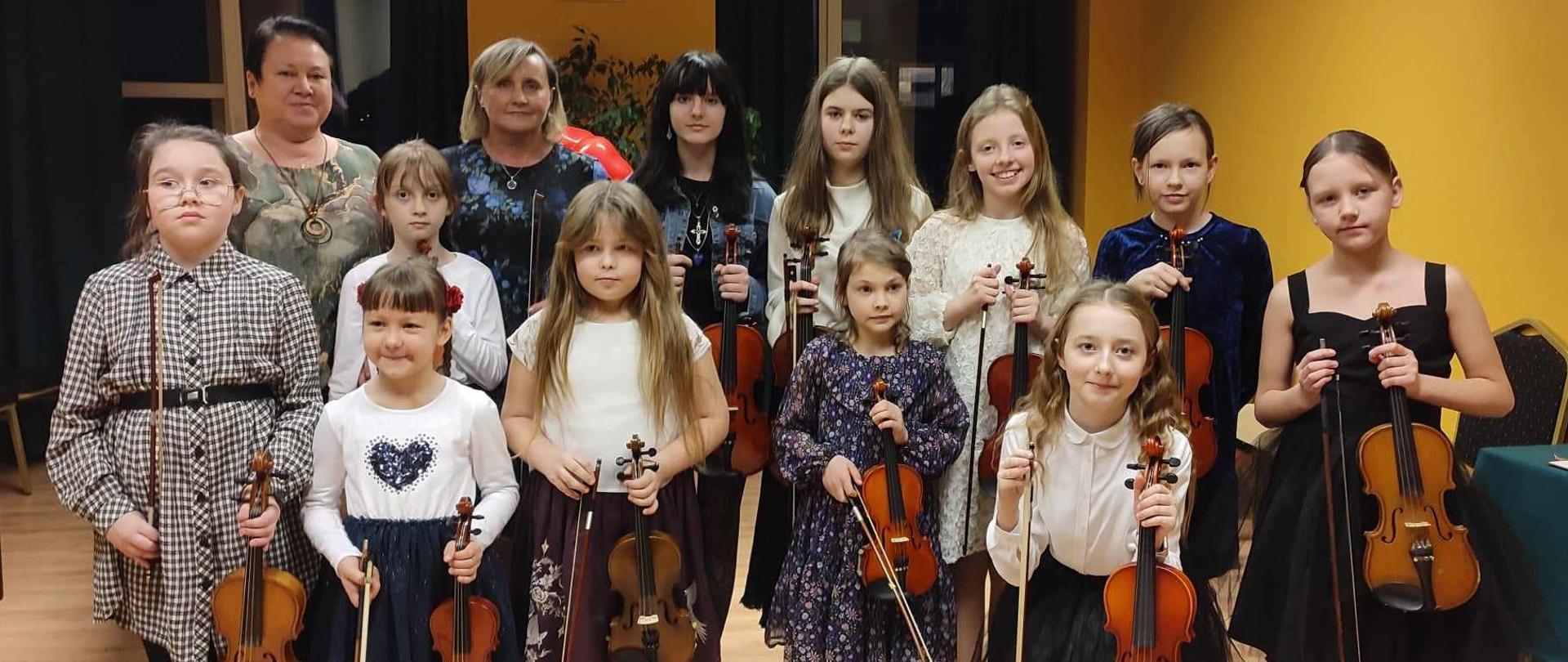 Zdjęcie grupowe uczniów klasy skrzypiec. Z przodu stoją niższe dziewczynki, z tyłu wyższe. Każda uczennica ma w rękach skrzypce i smyczek. z tyłu po lewej stronie obok siebie stoją dwie nauczycielki