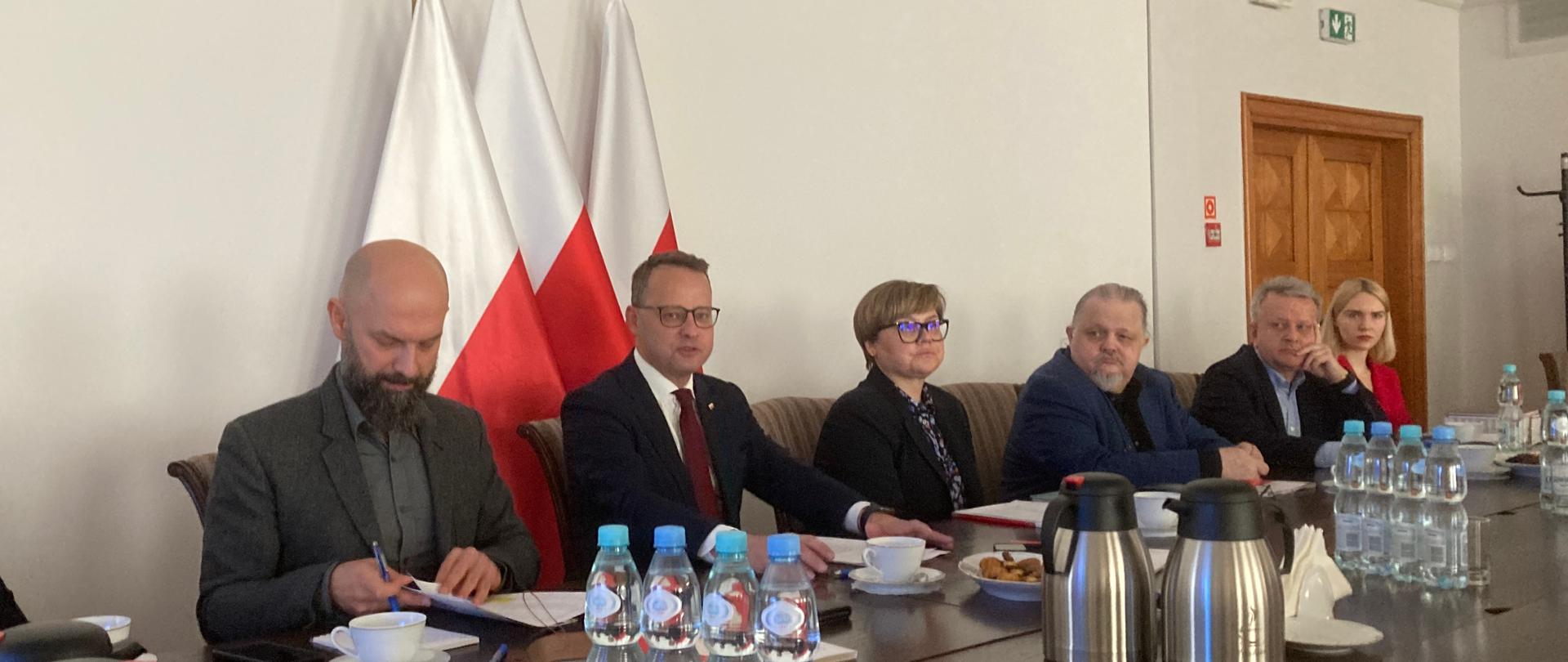 Grupa ludzi siedząca przy stole. Na drugim planie widoczne flagi.