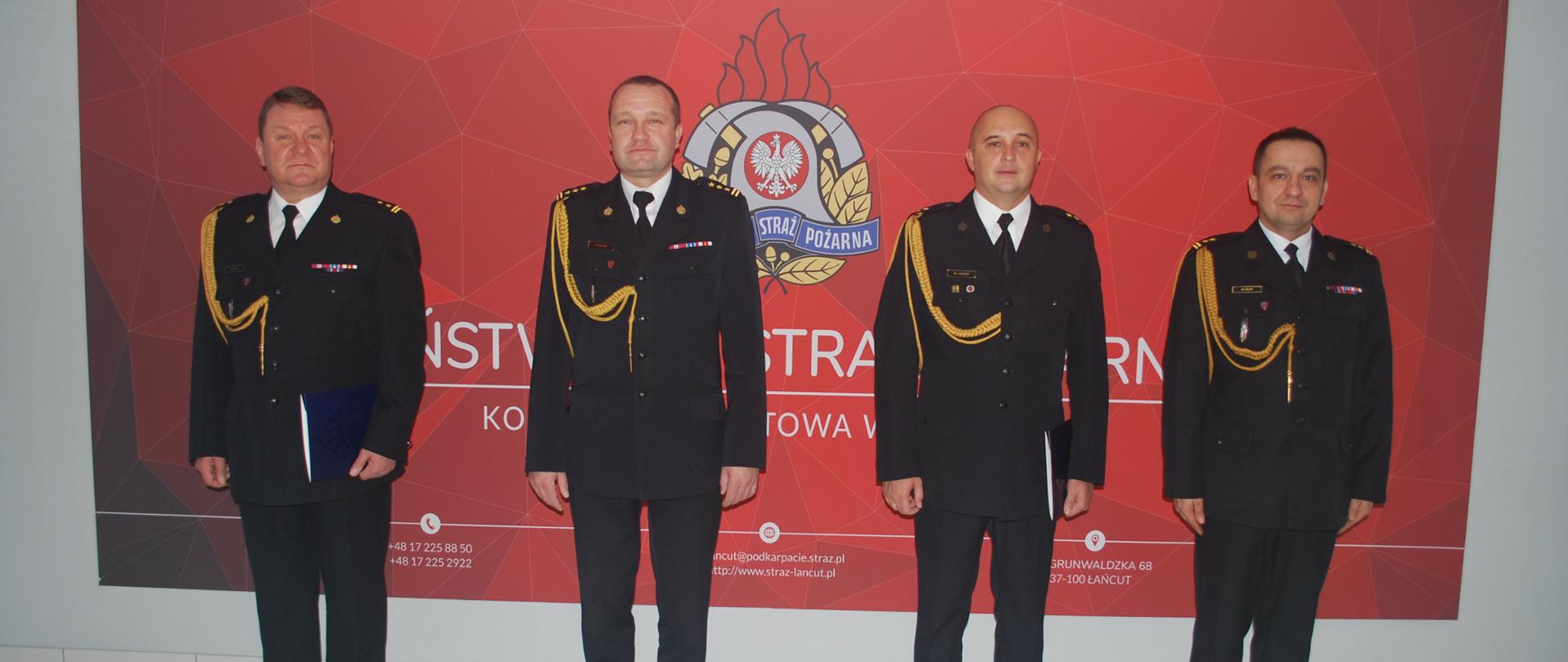 Zdjęcie przedstawia hol główny Komendy Powiatowej PSP w Łańcucie, na ścianie którego znajduje się tablica z logotypem Państwowej Straży Pożarnej. Na tle tablicy prezentują się w umundurowaniu galowym nominowani strażacy: brygadier Mariusz Kluz i młodszy kapitan Maciej Janusz w towarzystwie Komendanta Powiatowego Państwowej Straży Pożarnej starszego brygadiera Bogusława Goleni i Naczelnika Wydziału Organizacyjno-Kwatermistrzowskiego brygadiera Marcina Filipa.