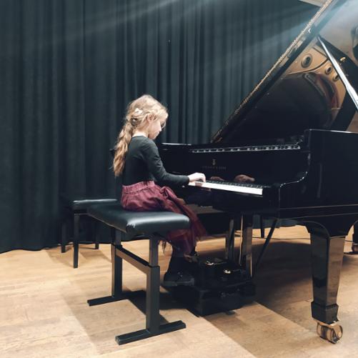 Dziewczynka gra na fortepianie, w tle czarna kurtyna.