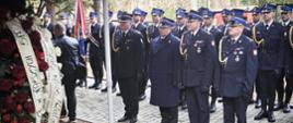 Strażacy w galowych mundurach biorą udział w ceremonii pogrzebowej na Cmentarzu Wojskowym w Przemyślu. Na pierwszym planie widoczne wieńce z biało-czarnymi wstęgami, w tle kompania honorowa i poczet sztandarowy PSP.