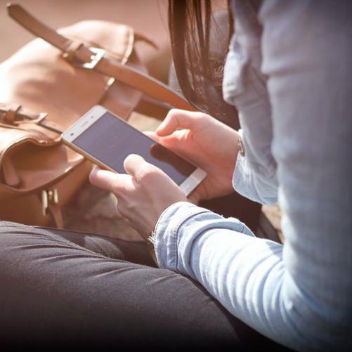 Kobieta siedzi po turecku i ogląda ekran smartfona, przed nią stoi torebka.