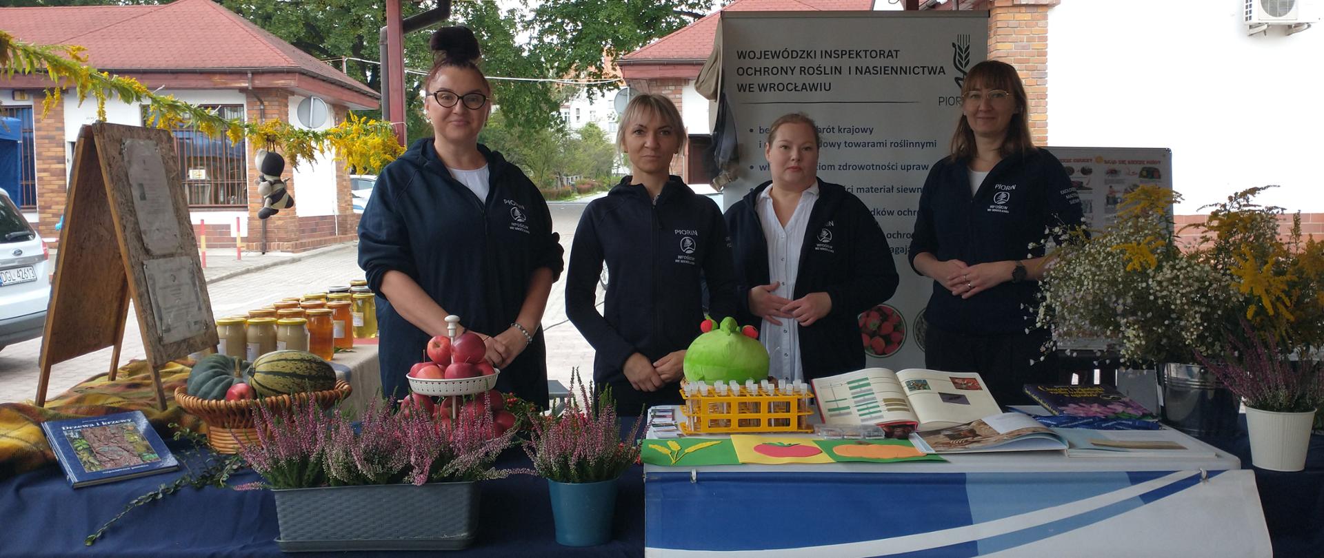 Na stoisku Wojewódzkiego Inspektoratu Ochrony Roślin i Nasiennictwa we Wrocławiu cztery kobiety prezentują materiały informacyjne oraz produkty ogrodnicze, m.in. warzywa, owoce, kwiaty i sadzonki. Na pierwszym planie widoczny jest baner z nazwą instytucji.
