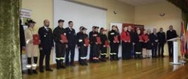 Wspólne zdjęcie strażaków zaproszonych gości oraz przedstawicieli Młodzieżowych Drużyn Pożarniczych którzy trzymają czerwone teczki.