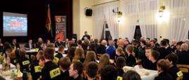 Widok boczny sali. Uczestnicy uroczystości siedzą przy stolikach. Na drugim przy wspólnym stole siedzą zaproszeni goście, jedna osoba stoi i przemawia do mikrofonu, w tle prezentacja 2023 MDP i OSP.