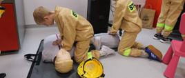 Dwoje dzieci ubranych w beżowe stroje strażackie ćwiczy udzielanie pierwszej pomocy na fantomie leżącym na podłodze sali edukacyjnej. Jedno z dzieci wykonuje uciski klatki piersiowej, a drugie klęczy obok i pomaga w ćwiczeniach. Obok fantomu leży żółty hełm strażacki. W tle widoczne są elementy wyposażenia sali dydaktycznej Państwowej Straży Pożarnej. Zdjęcie przedstawia praktyczną naukę zasad pierwszej pomocy prowadzoną w formie zajęć edukacyjnych dla dzieci.