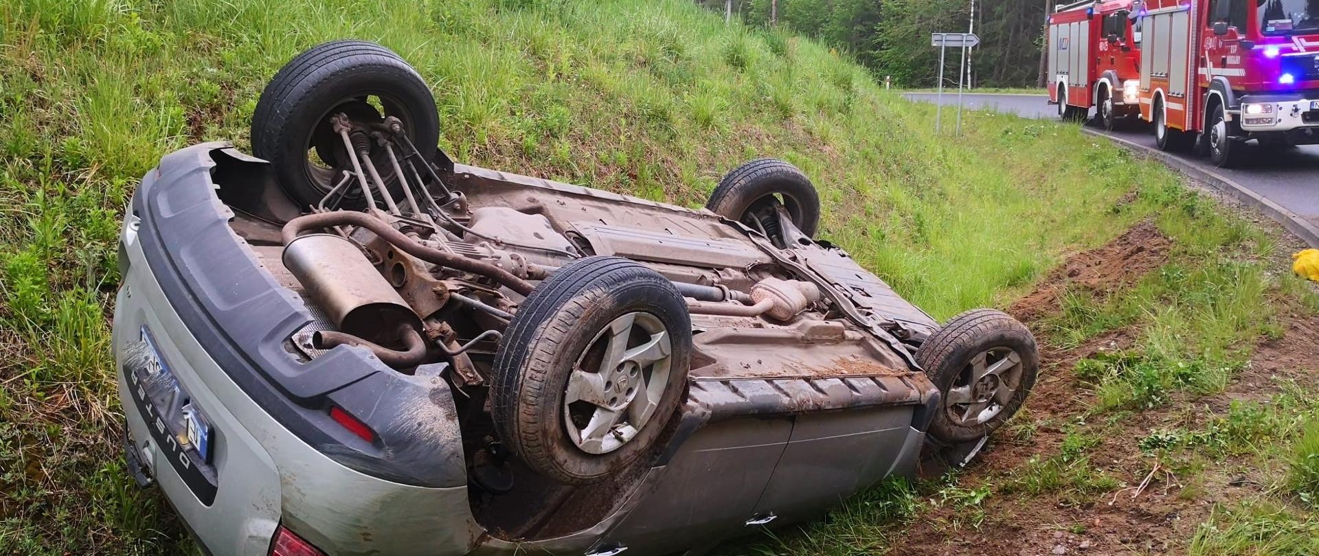 Zdjęcie przedstawia samochód w rowie kołami do góry. W drugim planie znajdują się pojazdy pożarnicze PSP