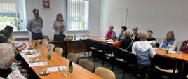 Kobieta i mężczyzna stojąc przemawiają na sali konferencyjnej. Obok przy stolikach siedzą inne kobiety. Na stole stoją komputer, szklanki, woda w butelkach oraz ciasto i cukierki. W tle baner reklamowy oraz godło Polski