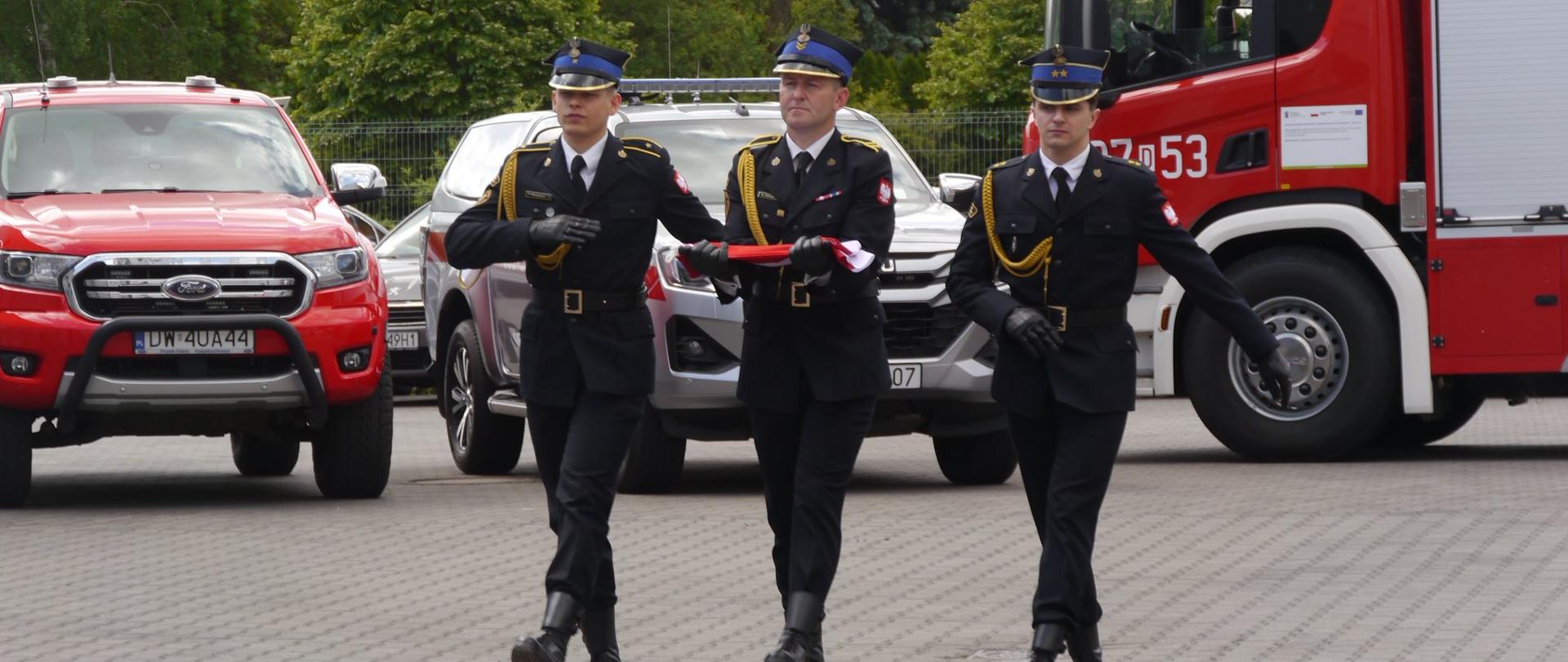 Strażacy pocztu flagowego maszerują z flagą Państwową, w tle znajdują się pojazdy pożarnicze