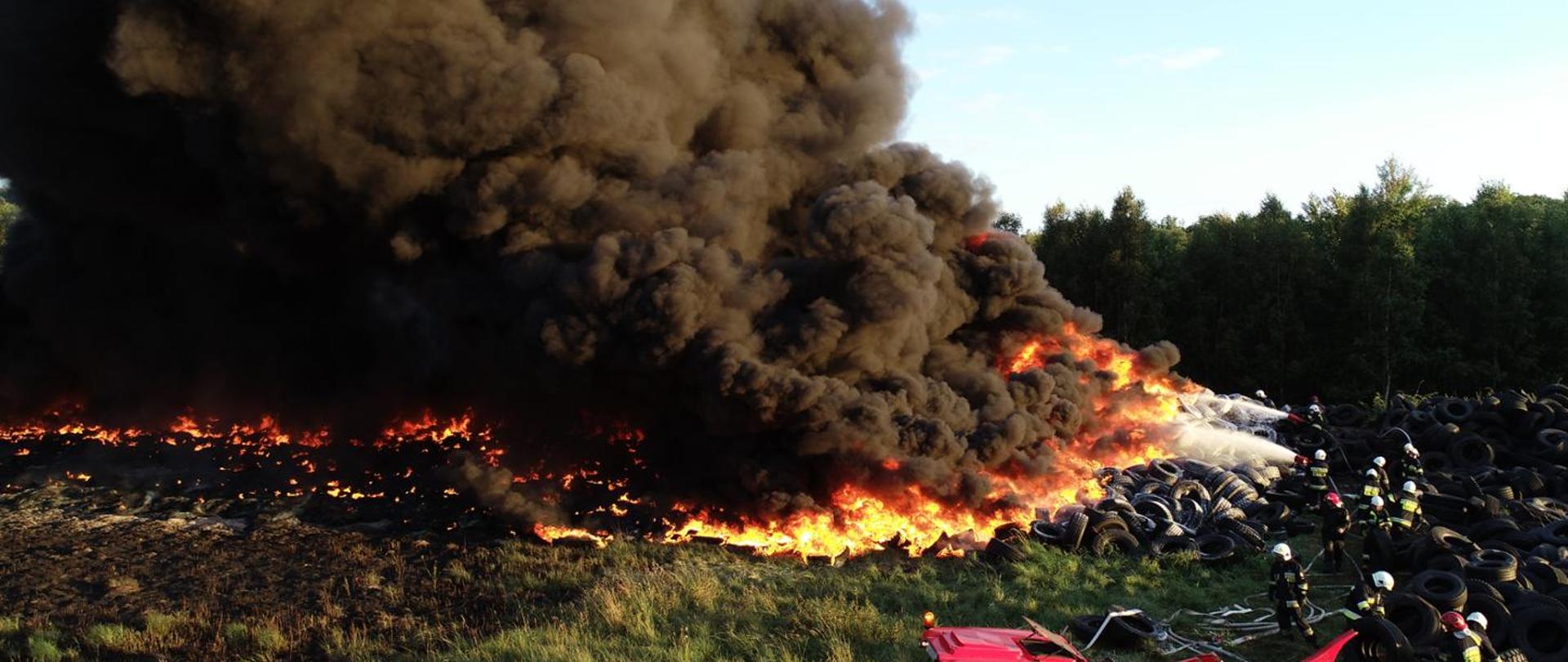 Fotografia przedstawia Płonące składowisko opon, w centralnej części zdjęcia widoczna hałda opon objęta płomieniami, nad pożarem unoszą się kłęby gęstego ciemnego dymu. Z prawej strony fotografii kilkunastu strażaków podaje prądy piany średniej na front pożaru, broniąc przed rozprzestrzenieniem się pożaru na resztę składowiska, dalej za składowiskiem widoczny las.