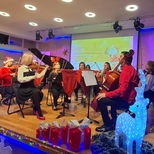 Zdjęcie przedstawia zespół smyczkowy podczas wykonania utworu na scenie w sali koncertowej, widoczne dekoracje świąteczne, w tle otwarte fortepian oraz ekran multimedialny