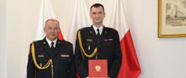 Dwóch funkcjonariuszy w mundurach wyjściowych ze sznurem stoi obok siebie jeden z nich trzyma czerwoną teczkę za nimi znajdują się trzy flagi Polski na ścianie wisi obraz.