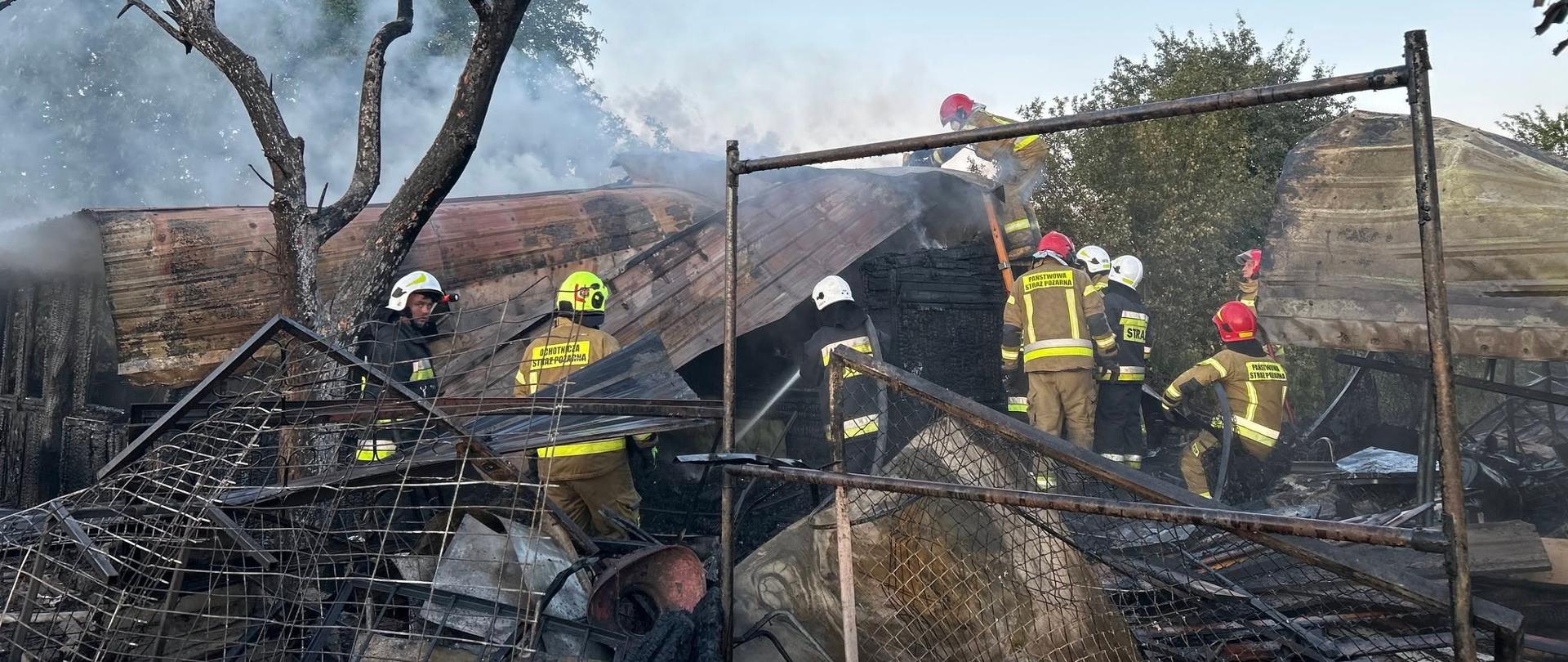 Na zdjęciu widać kilku strażaków pracujących przy dogaszaniu budynku. Znajdują się oni na terenie mocno zniszczonym przez pożar, wśród zwęglonych elementów konstrukcji, metalowych prętów, blach i gruzu. Strażacy pracują na pozostałościach dachu lub konstrukcji budynku. Nad pogorzeliskiem unosi się dym.

