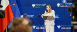 Minister funduszy i polityki regionalnej Katarzyna Pełczyńska-Nałęcz stoi przy mównicy i przemawia do mikrofonu. Po jej prawej stronie flagi Polski i UE, za jej plecami granatowy baner z logotypami MFiPR