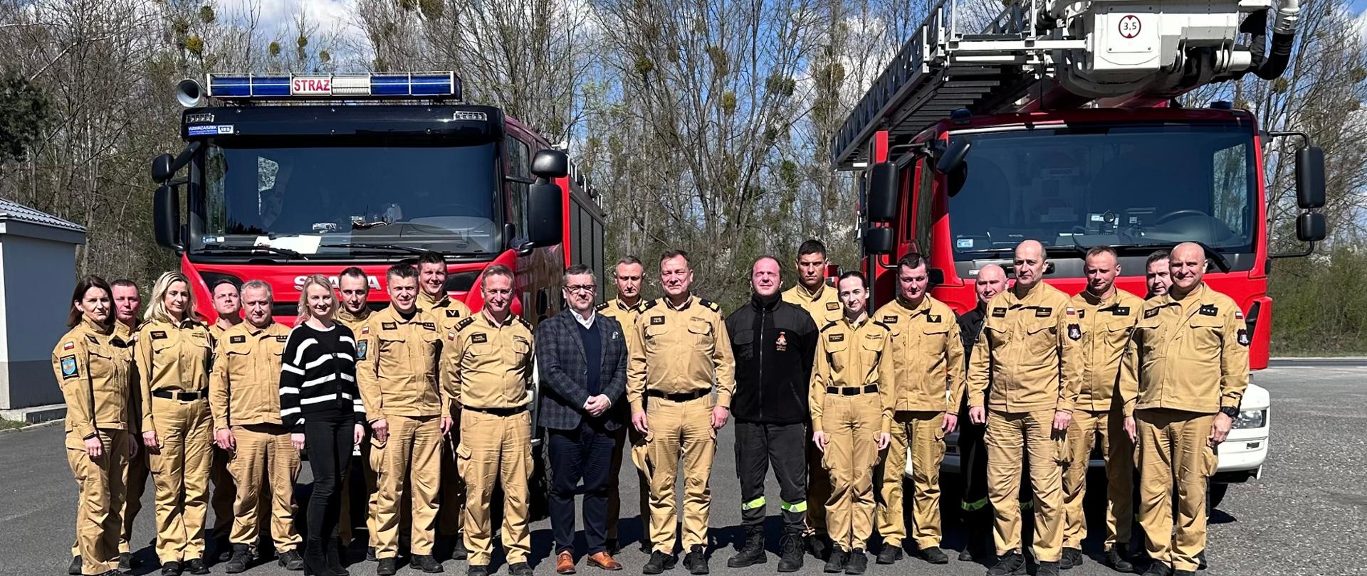 Na zdjęciu znajdują się strażacy w umundurowaniu służbowym ustawieni do zdjęcia na tle samochodów bojowych