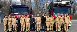 Na zdjęciu znajdują się strażacy w umundurowaniu służbowym ustawieni do zdjęcia na tle samochodów bojowych
