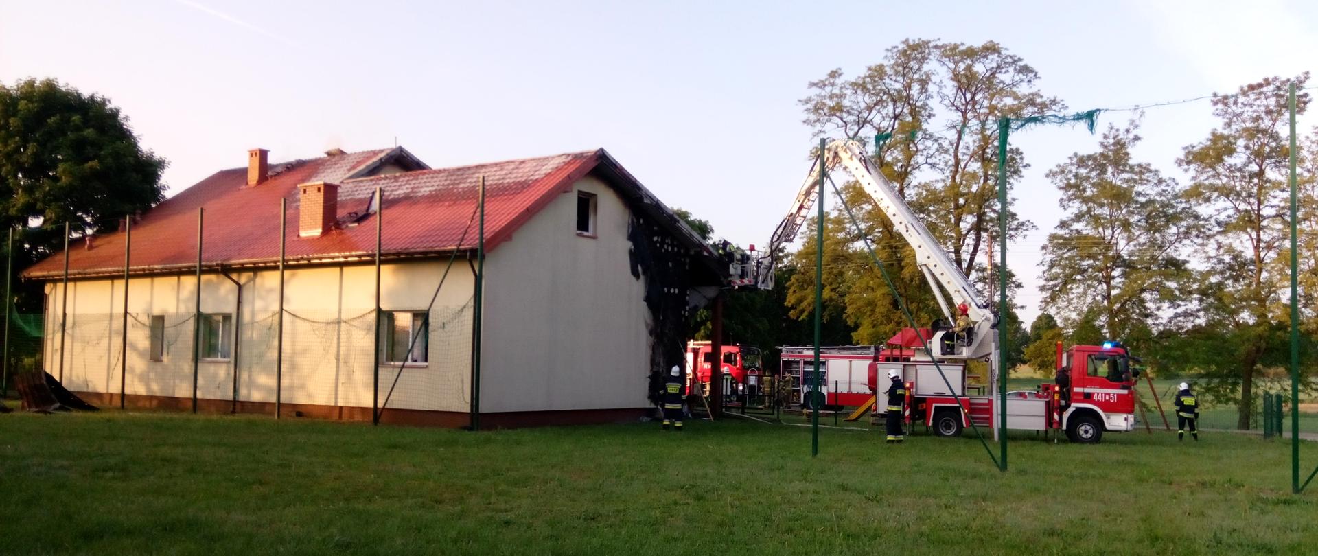 Zdjęcie przedstawia część zniszczonej elewacji budynku remizy. Obiekt w kolorze żółtym, dach w kolorze czerwonym. Obok budynku widoczne są samochody strażackie w kolorze czerwonym oraz pracujący strażacy.