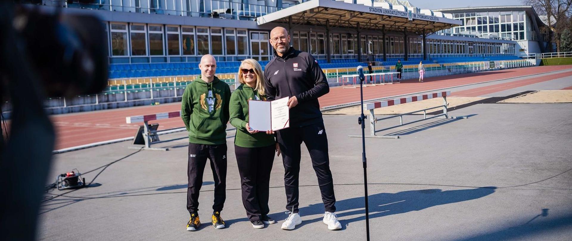 Minister sportu i turystyki Jakub Rutnicki stoi przed stadionem lekkoatletycznym AWF Warszawa i prezentuje decyzję o dofinansowaniu jego remontu. Obok niego dwie osoby w zielonych bluzach AWF
